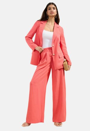 Kobieta nosząca koralowe spodnie o szerokich nogawkach i dopasowaną marynarkę na białej bluzce, trzyma małą wiklinową torebkę.