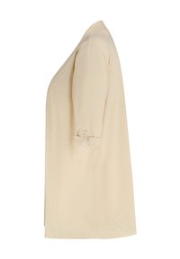 Blouse beige à manches courtes avec une coupe ample, présentant un détail de manche retroussée avec bouton et une texture lisse et légère.