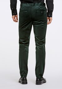 Pantaloni verde velluto con una texture liscia, dotati di due tasche posteriori con bottoni e una vestibilità su misura, abbinati a scarpe nere.