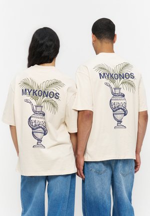 Két ember bézs színű, túlméretezett pólót visel, melyen kék kígyóval körbetekert váza és zöld pálmalevelek láthatók, hátul felirat: „MYKONOS”.