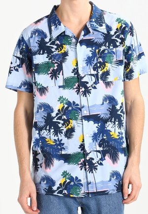 Koszula na krótki rękaw z nadrukiem w niebieskie tropikalne kwiaty i palmy, z dwoma kieszeniami na klatce piersiowej, noszona przez osobę w niebieskich dżinsach.