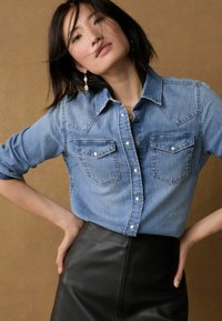 Hellblaues Denim-Hemd mit Knöpfen, zwei Brusttaschen und spitzem Kragen, kombiniert mit einem schwarzen Leder-Bleistiftrock.