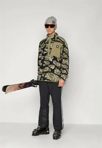 Πολυεστερική μπλούζα σε καμουφλάζ πράσινου και μαύρου χρώματος, με φερμουάρ στον γιακά και τσέπη στο στήθος. Μαύρο παντελόνι με λευκές ρίγες; Φοράει σκιέρ μποτάκια και γυαλιά ηλίου.