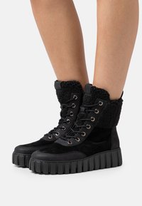 Botas negras con cordones que presentan un upper de ante, un cuello peludo y una suela de goma gruesa y estriada con ojales metálicos.