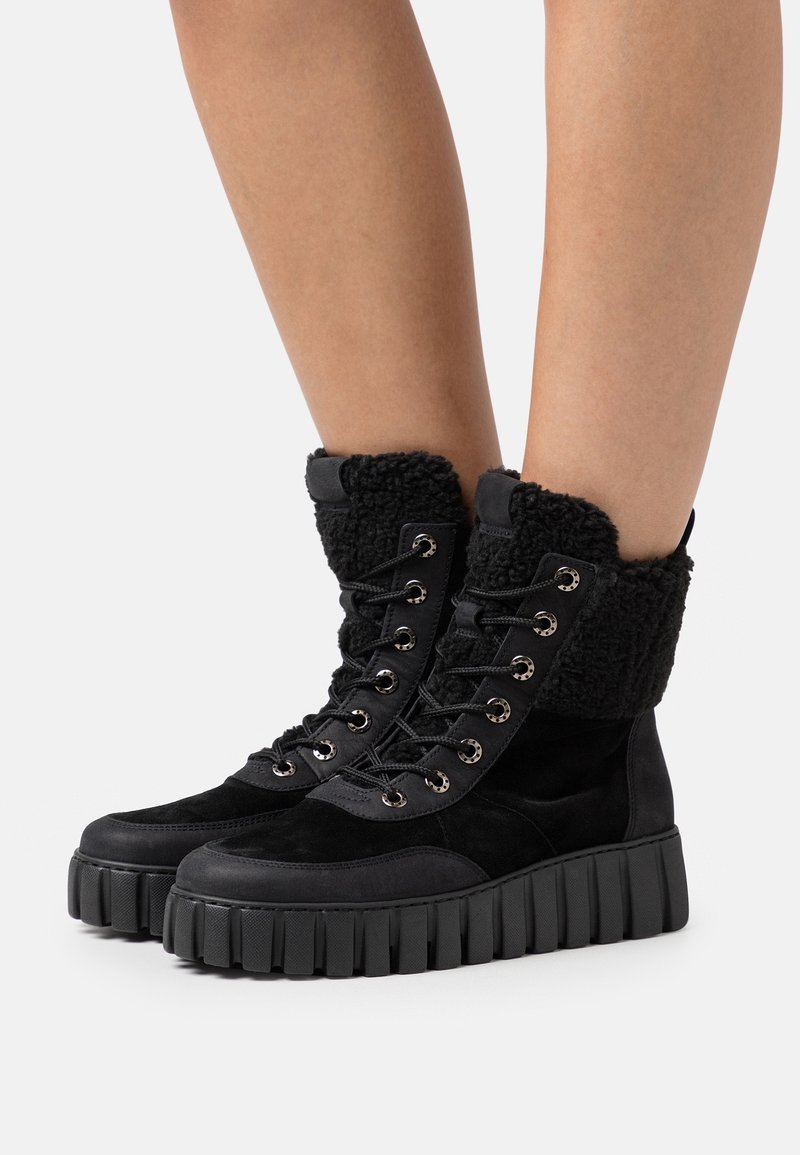 Botas negras con cordones que presentan un upper de ante, un cuello peludo y una suela de goma gruesa y estriada con ojales metálicos.