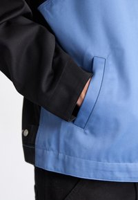 Tofarvet jakke med sort ærme og blå krop, med en glat tekstur og detaljer med sidelomme. Synlige metalknapper.