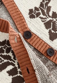 Close-up of knitted fabric with brown floral patterns, rust-colored ribbed edges, buttonholes, and dark brown buttons.
