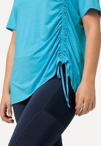 Hellblauses Sporttop mit seitlichem Raffung und Kordelzügen, kombiniert mit marineblauen Leggings, die über strukturierte Mesh-Einsätze verfügen.