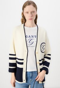 Cardigan crem cu dungi navy, guler rotund și nasturi aurii. Purtați peste un tricou alb cu text și logo bleu. Croială casual.
