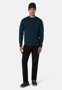 Dunkeltürkises Sweatshirt mit Rundhalsausschnitt, schwarze Hosen und dunkelblaue Sneakers. Das Outfit wird durch eine passende Mütze ergänzt. Einfaches Design.