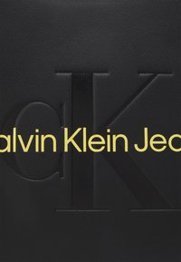 Black textured leather bag featuring an embossed logo design with "Calvin Klein Jeans" in bold yellow letters.