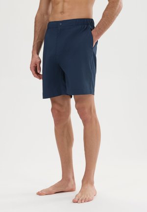 Homme portant un short bleu marine arrivant aux genoux avec une taille élastique et un bouton, debout pieds nus, une main dans la poche.