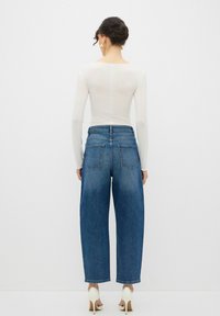 Top blanco de manga larga combinado con jeans de denim azul de pierna ancha, que cuentan con dos bolsillos traseros y un acabado limpio, usados con tacones color blanco sucio.