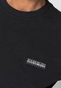 Svart bomullströja med rund halsringning och en rektangulär logotypremse som står "NAPAPIJRI geographic" i vitt på vänster bröst.