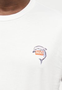 Κοντινή λήψη ενός λευκού T-shirt με ένα μικρό γραφικό δελφινιού και πορτοκαλί λογότυπο «DIESEL» στο στήθος.