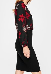 Femme portant un chemisier à manches longues noir avec un imprimé floral rouge et blanc, rentré dans une jupe noire taille haute longueur genou.