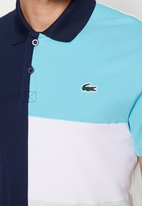 Męska koszulka polo z blokami kolorów w granacie, jasnym niebieskim, białym i miętowej zieleni. Posiada kołnierzyk, dwa guziki i ozdobę z logo krokodyla.