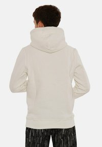 Cream-colored hoodie with a large, soft hood and ribbed hem. Made of a smooth fabric with a relaxed fit, worn with patterned dark pants.