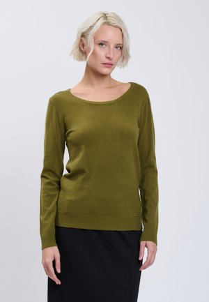 Kvinde med kort blondt hår iført en olivengrøn langærmet sweater og en sort nederdel, stående mod en ensfarvet baggrund.