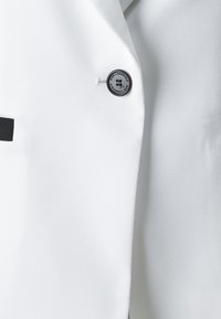 Biely blazer s hladkou textúrou látky, ostrými klopami a čiernym logom na gombíku umiestneným na prednej ľavej strane.