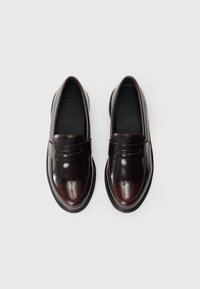 Mocassini color bordeaux con finitura patinata lucida, caratterizzati da un tacco basso, punta tonda e un dettaglio con cinturino frontale. Soletta nera testurizzata.