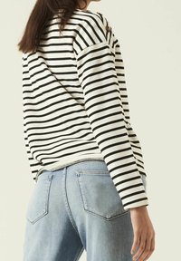 Randig långärmad topp i krämvit med svarta horisontella ränder, med en knappdetalj på axeln. Matchad med ljusblå denimjeans.