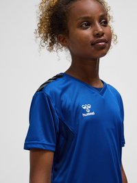 Młoda osoba z kręconymi włosami w niebieskiej sportowej koszulce z logo "hummel", lekko patrząca w bok na jednolitym jasnym tle.