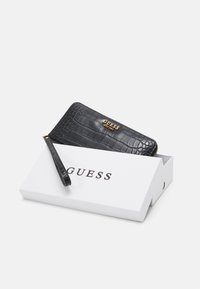 Sort faux krokodilleder pung med et guld Guess-logo, lynlås lukning og aftagelig håndledsrem, ligger i en hvid mærket æske.