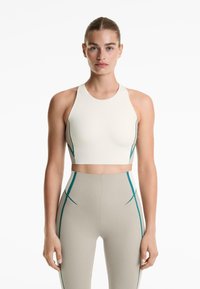 Kvinna som står framåtklädd i en krämfärgad ärmlös crop top och högmidjade beigefärgade leggings med blå vertikala randdetaljer.