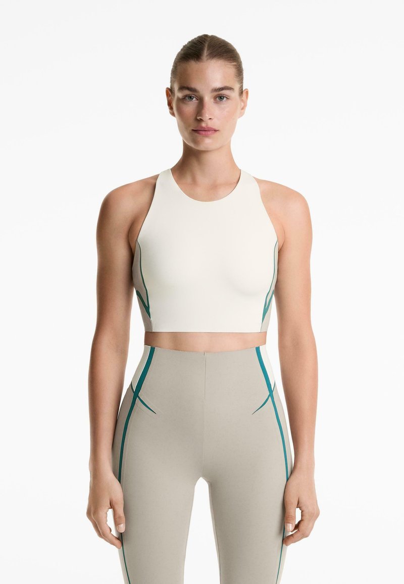Kvinna som står framåtklädd i en krämfärgad ärmlös crop top och högmidjade beigefärgade leggings med blå vertikala randdetaljer.