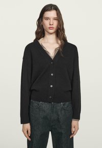 Czarny kardigan z dekoltem w serek, długimi rękawami, pięcioma guzikami z przodu oraz ściągaczami na mankietach. Noszony na paskowanej bluzce i zestawiony z ciemnymi dżinsami.