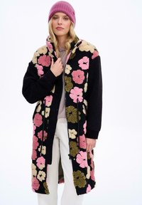 Čierny, plyšový kabát s mnohofarebn�ými kvetinovými vzormi v ružovej, krémovej a zelenej farbe; má zips a vysoký golier, nosený cez svetlý sveter.