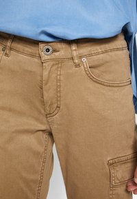 Beżowe bawełniane spodnie typu cargo z frontowym zapięciem na guziki, bocznymi kieszeniami i detalami szwów. Tkanina ma gładką fakturę i delikatny połysk.