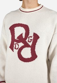 Creme sweater med tekstureret strik. Har et stort burgunderfarvet grafisk "RDG" logo og en burgunderfarvet krave med kant. Blødt og komfortabelt materiale.