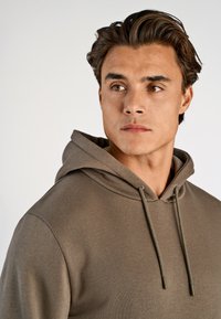Sudadera color verde oliva en un tejido suave y texturizado. Cuenta con capucha ajustable y un corte relajado. Costuras suaves y detalles de diseño minimalistas.