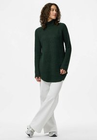 Donna con capelli ricci che indossa un maglione a collo alto verde scuro, pantaloni bianchi a gamba larga e sneakers argento, in piedi contro uno sfondo neutro.
