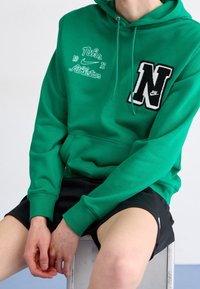 Person som bär en grön Nike-hoodie med broderade logotyper och svarta träningsshorts, sittande på en vit pall mot en enfärgad bakgrund.