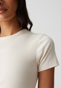 T-shirt crema a costine con scollatura rotonda e maniche corte, caratterizzato da una texture morbida e una forma aderente, che mette in evidenza le spalle e la parte superiore del petto.