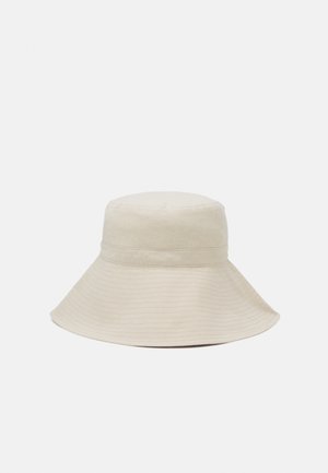 Beżowy kapelusz typu bucket z szerokim rondem i detalem przeszywanego paska, przedstawiony na jednolitym jasnoszarym tle.