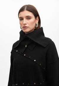 Suéter de punto negro con un cuello alto y acentos de botones dorados. Presenta un cuello acanalado texturizado y tachuelas doradas horizontales en la parte delantera.