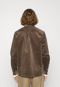 Camisa de pana marrón con canalones verticales, puños abotonados y un yugo trasero. Presenta un cuello clásico y una textura suave.