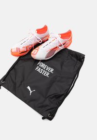 Puma fotbollsskor i vitt och orange med ett nätmönster, ihopparade med en svart dragsnörpåse med vit text och logotyp.