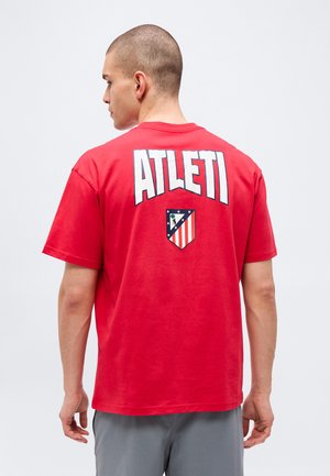 Homme vu de dos portant un t-shirt rouge avec "ATLETI" et un logo en forme de bouclier à rayures rouges, blanches et bleues dans le dos.
