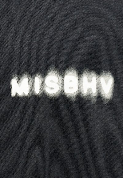 Μαύρο ύφασμα με λευκά γράμματα σε σχέδιο στένσιλ "MISBHV" που εμφανίζονται ελαφρώς θολά ή ξεθωριασμένα στις άκρες.