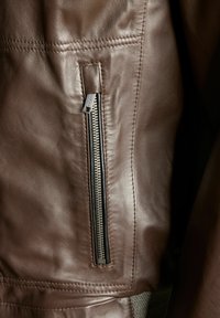 Detail van een bruin leren jack met een ritszak aan de linkerkant. Gladde textuur met zichtbare stitching en een blootgestelde metalen rits.