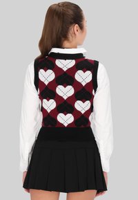 Vesta tricotată în nuanțe de burgundy, negru și alb, cu un model de inimioare; guler și tiv cu reazem; asortată cu o cămașă albă cu guler și o fustă plisată neagră.