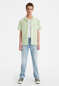 Joven de pie vistiendo una camisa de rayas amarillas y azules de manga corta, una camiseta blanca, jeans de mezclilla claros y zapatillas negras, sobre un fondo blanco.