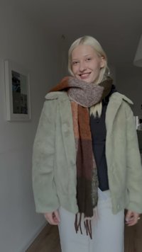 Manteau en fausse fourrure vert clair avec larges revers, associé à une écharpe multicolore aux nuances de brun, crème et rouille. Pantalon blanc.