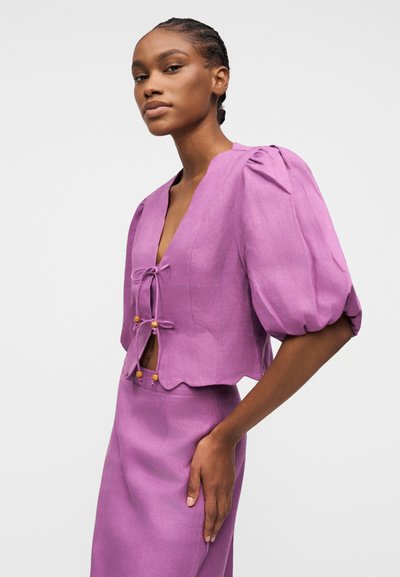 Femme aux cheveux tressés portant un haut violet à manches bouffantes avec des fermetures à nouer à l'avant, assorti à une jupe, sur un fond uni.