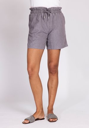 Person trägt graue Shorts mit Kordelzug und ein weißes Oberteil sowie schwarz-weiße, geflochtene Sandalen mit offenen Zehen an nackten Füßen.